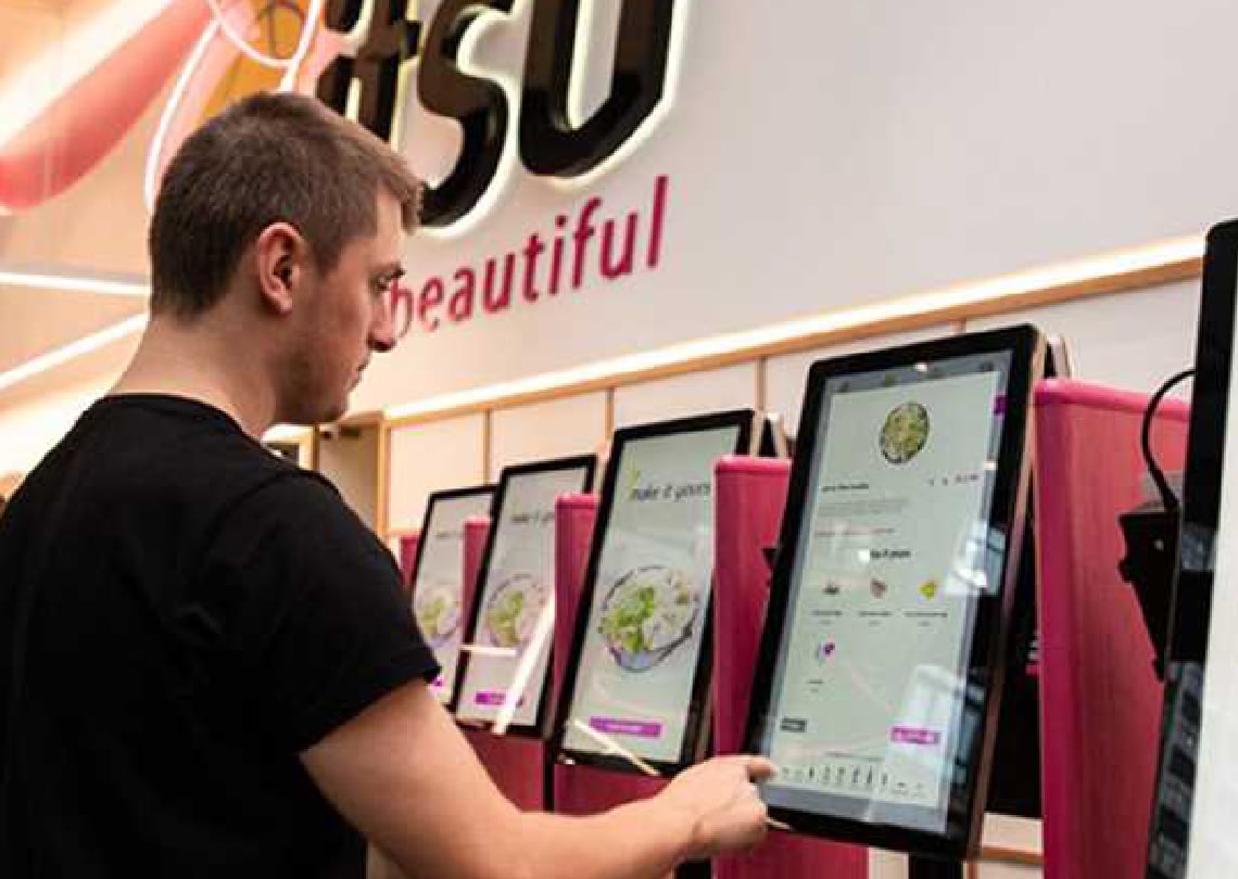 Homem interagindo com quiosques digitais em um ambiente moderno, com a marca 'it’s so beautiful' ao fundo, ideal para uma experiência de compra inovadora.