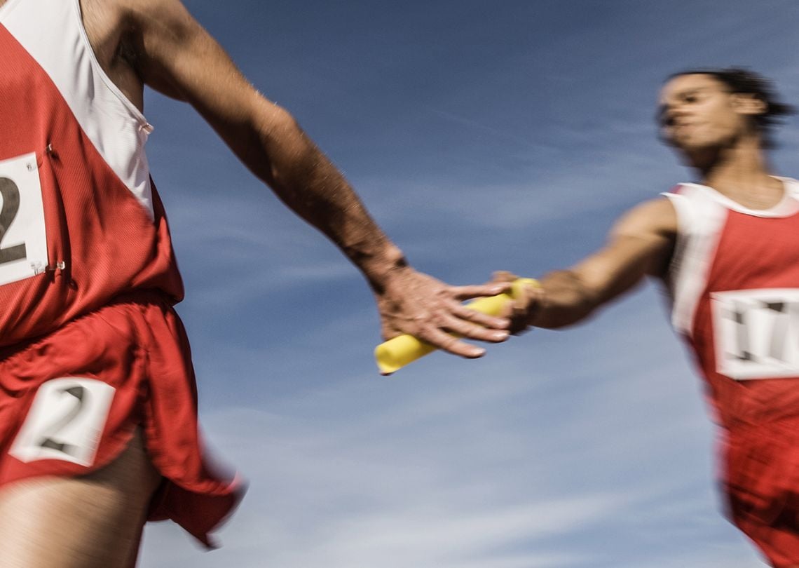 Atletas competindo em uma corrida de revezamento, um corredor passa o bastão para o outro sob um céu azul, simbolizando velocidade e trabalho em equipe.