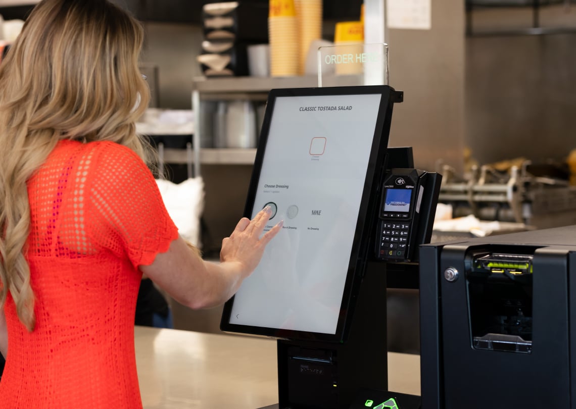 Imagem de uma mulher utilizando um quiosque de autoatendimento para pedir uma salada tostada em um restaurante moderno. A tecnologia facilita o pedido personalizado.