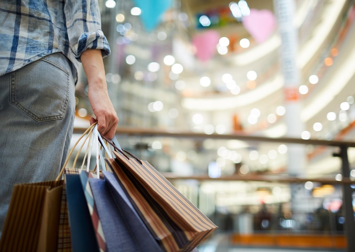 Pessoa segurando sacolas de compras em um shopping moderno, com muitas luzes e um ambiente vibrante ao fundo.