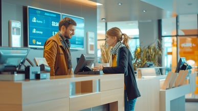 Atendente e cliente conversando em um balcão de check-in, com telas informando voos e check-in auto.