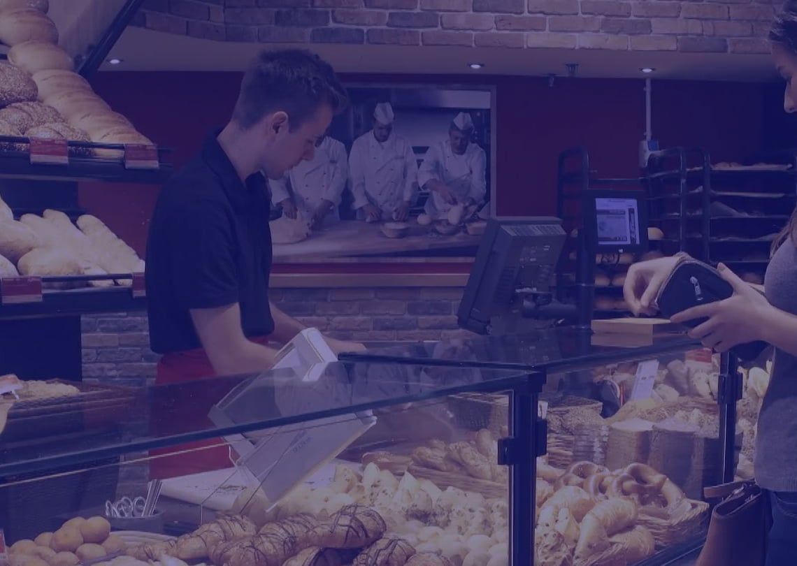 Imagem de uma padaria com diversas opções de pães, onde um atendente serve uma cliente que consulta o menu. Uma variedade de pães é exibida em uma vitrine atraente, destacando a qualidade e frescor dos produtos.