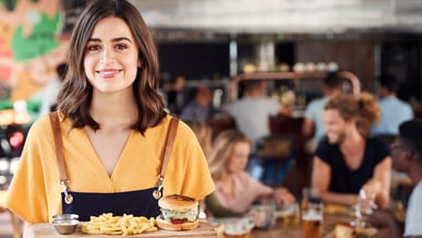 Imagem de uma garçonete servindo um prato com hambúrguer e batatas fritas em um restaurante moderno e animado, com clientes ao fundo.
