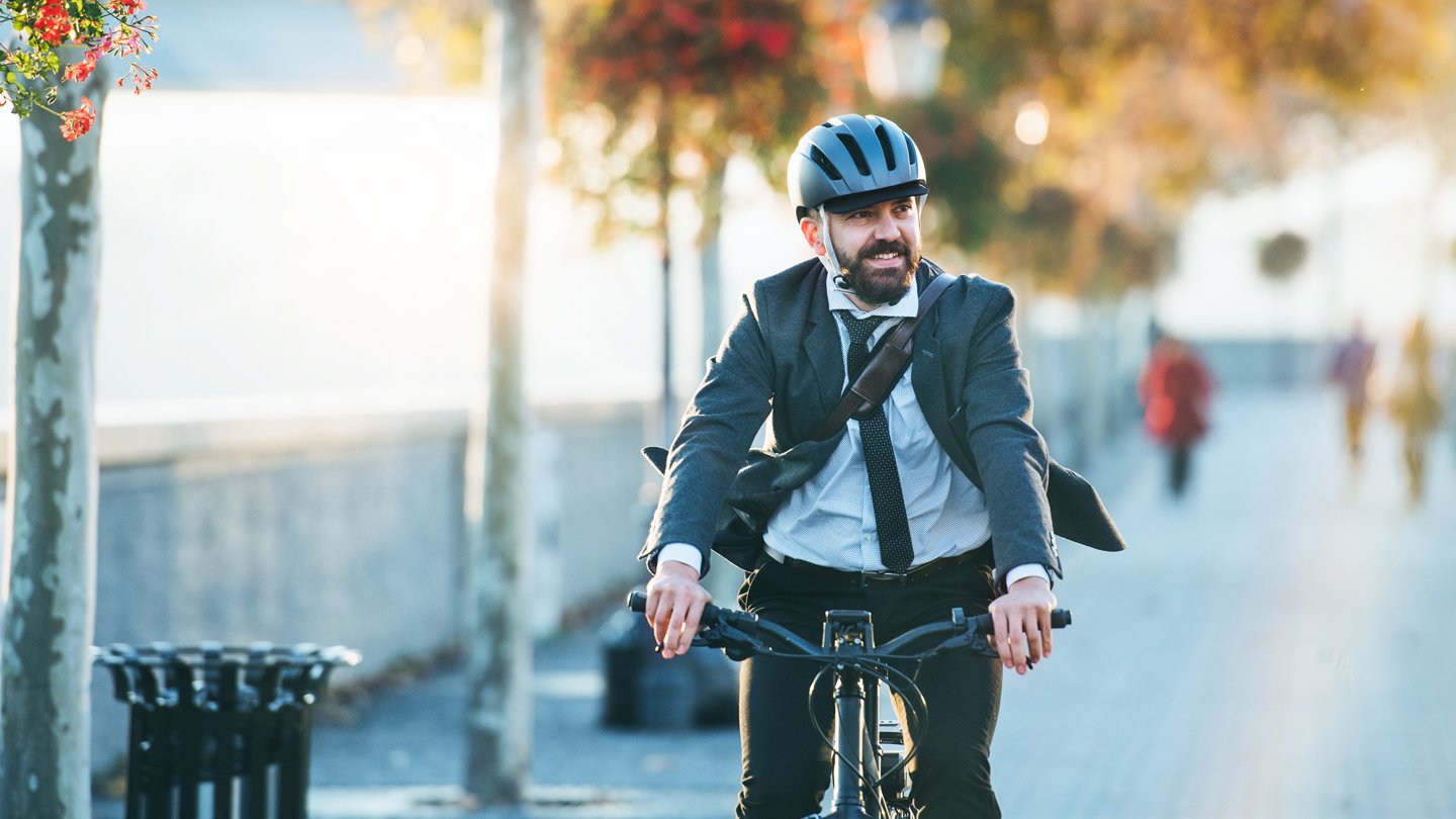 Homem de terno e camisa, usando capacete de ciclismo, pedalando de bicicleta em uma rua arborizada durante o dia com clima de outono, demonstrando mobilidade urbana sustentável.
