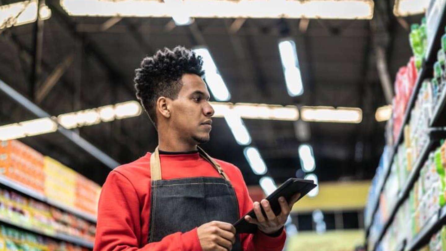 Um jovem trabalhador em um supermercado verifica produtos em uma prancheta, demonstrando foco e dedicação ao seu trabalho. Imagem que reflete o cotidiano do varejo.