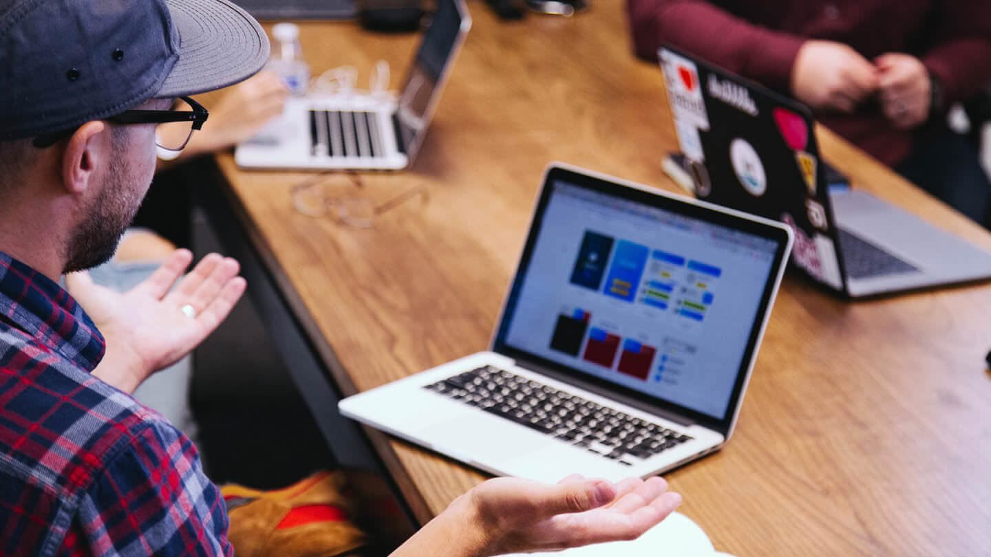 Imagem de uma reunião de equipe onde profissionais discutem projetos, utilizando laptops e gestos para troca de ideias. O foco está na colaboração e criatividade em um ambiente de trabalho moderno.