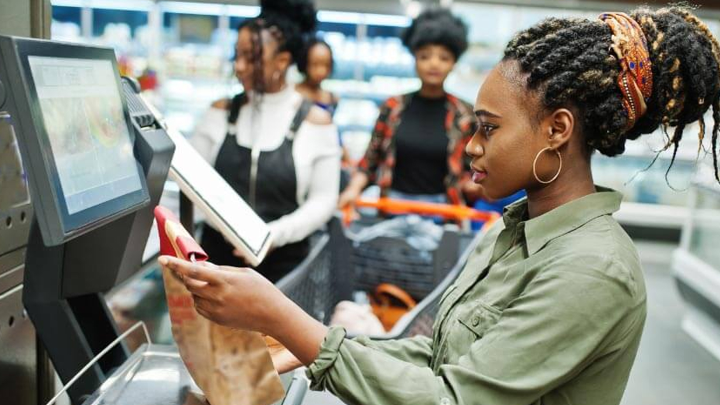 Mulher usando caixa de autoatendimento em supermercado, representando o uso de tecnologia nas compras. Ambiente de supermercado moderno com clientes ao fundo.