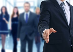 Homem de terno estendendo a mão em cumprimento, simbolizando networking e relações profissionais, com pessoas ao fundo em ambiente corporativo.