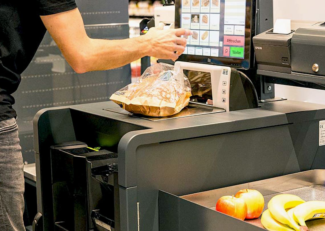 Imagem de um caixa de supermercado onde uma pessoa está escaneando produtos. O caixa digital mostra opções de itens. Frutas como maçãs e bananas estão visíveis no balcão.