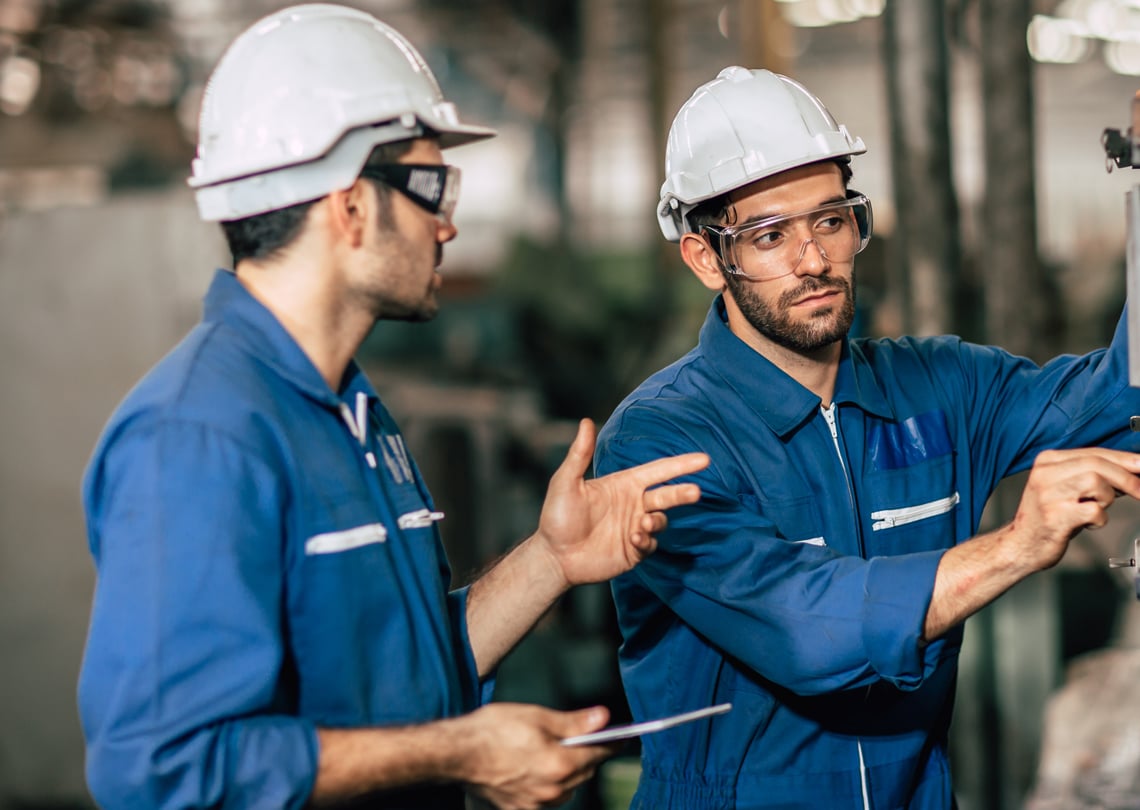 Dois engenheiros trabalhando em uma fábrica, usando equipamentos de proteção como capacetes e óculos de segurança, realizando inspeção técnica.