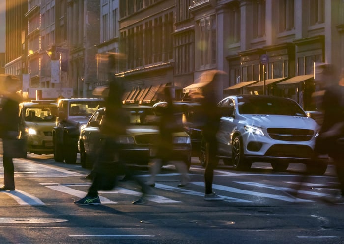 Imagem de pessoas atravessando a rua movimentada à noite na cidade, com carros estacionados e edifícios ao fundo, iluminação urbana e movimento.