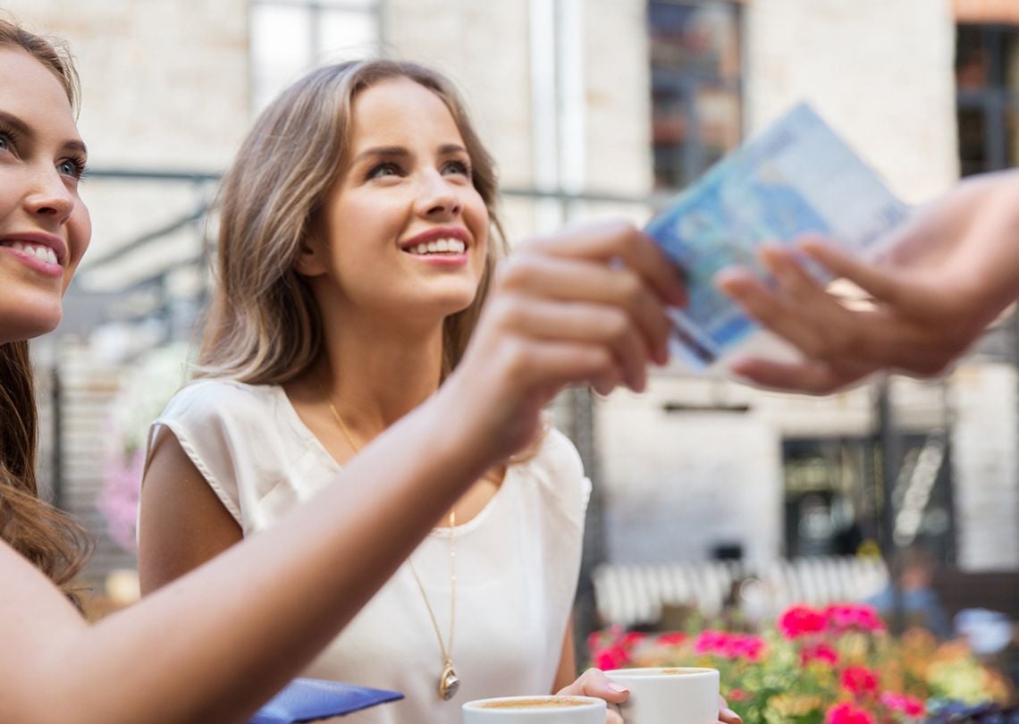 Duas mulheres sorrindo enquanto uma delas recebe um pagamento em dinheiro, rodeadas por flores e bebidas em um ambiente ao ar livre.