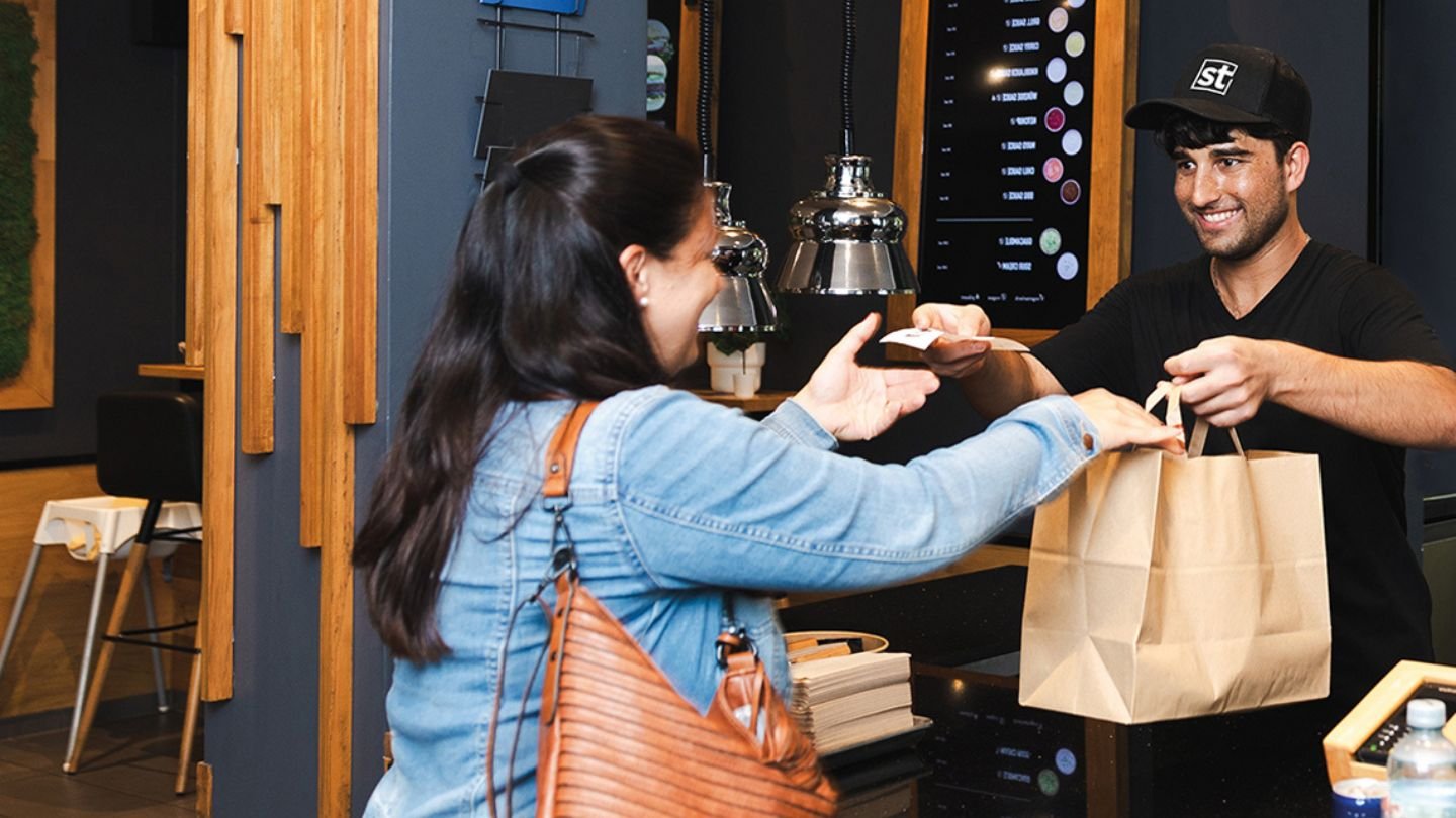 Cliente retirando pedido em cafeteria. Funcionário sorridente entrega sacola com café enquanto a cliente faz pagamento com cartão. Experiência de compra agradável.